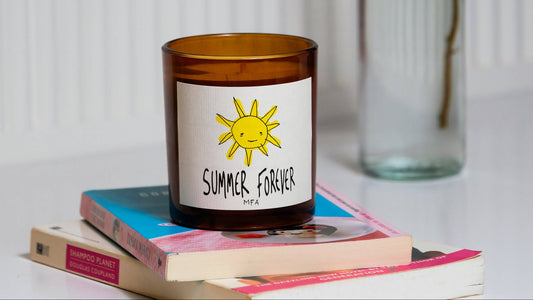 An amber glass candle jar with a white label featuring a yellow sun and the text 'SUMMER, FOREVER' and 'PFAA', styled on top of stacked books with a blurred glass bottle in the background.