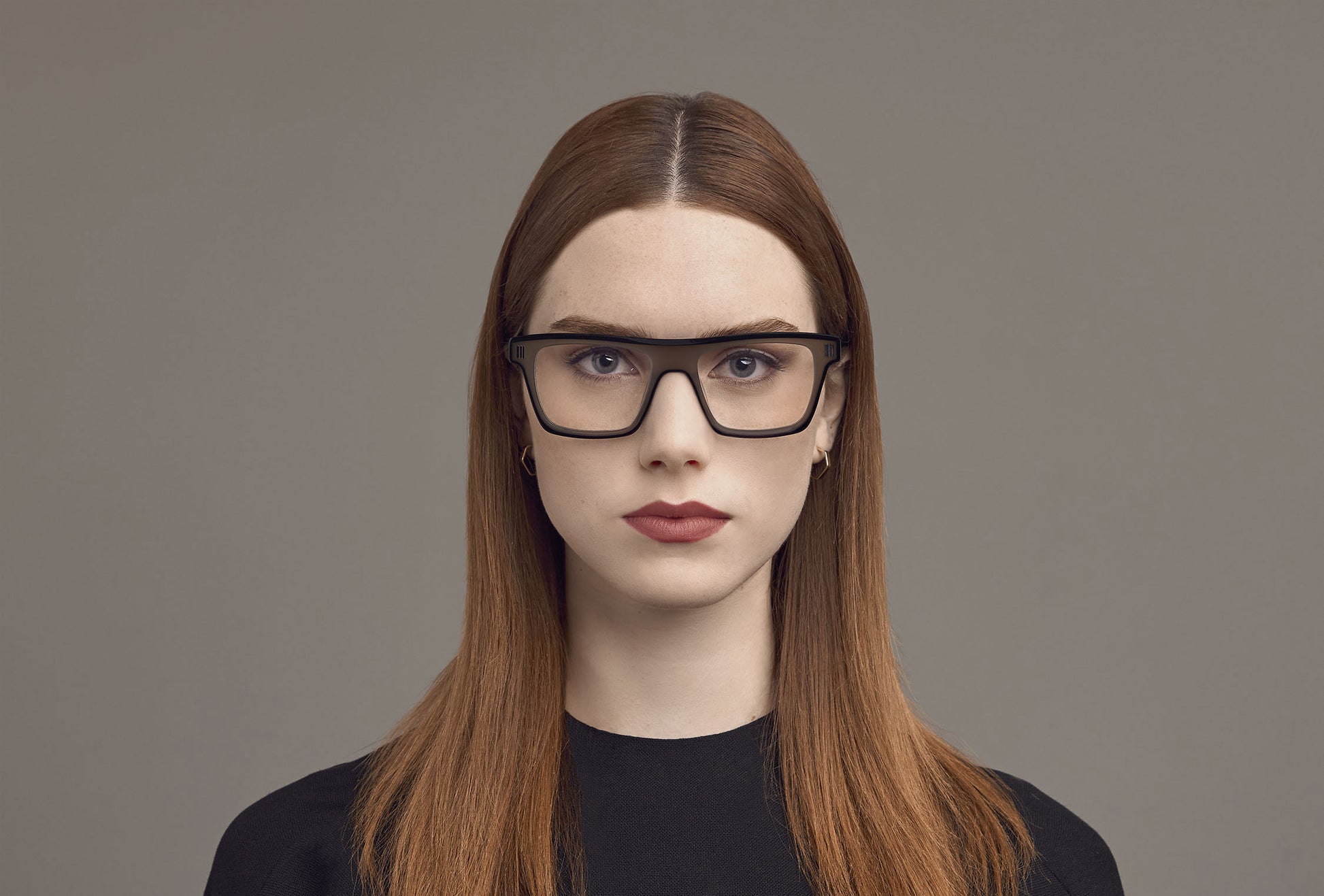 A woman with long auburn hair wearing black rectangular eyeglasses, photographed against a plain taupe-gray studio background.